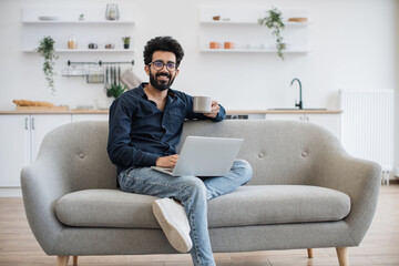 Portrait of joyful young adult in cozy clothes posing with drink in cup and portable computer in modern kitchen. Delighted arabian man in spectacles networking on couch using smart gadget at home.