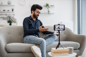 Serious arabian man in casual clothes sitting on grey sofa and communicating via video call on wireless laptop while recording video on modern smartphone. Concept of people and social media.