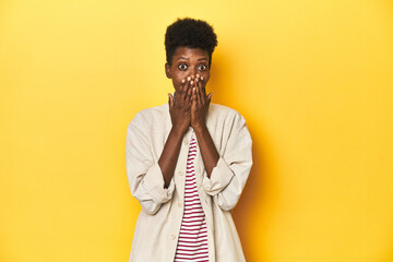 African woman, beige shirt, red striped tee, yellow studio, shocked, covering mouth with hands,...