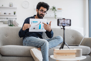 Young arabian financier with black beard filming himself on cell phone which fixed on tripod and showing financial statistics to subscribers in living room. Concept of technology and blogging.