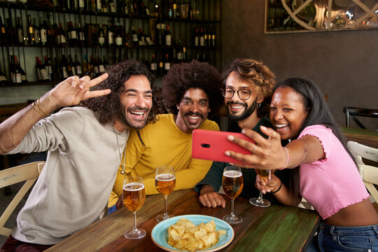 Multiracial gathering of friends celebrating in a bar and having fun while they taking a selfie of the group of people. Young and carefree they drink beer and eat while they take picture with a phone