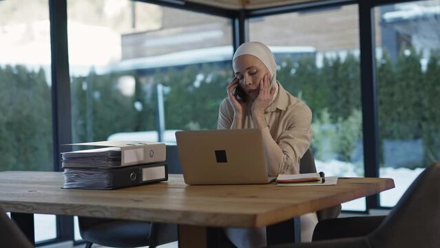 Cute female in hijab is sitting at the table in front of a laptop