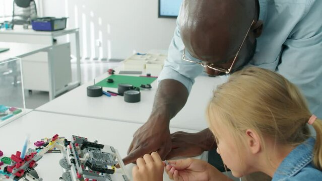 Over-shoulder arc shot of colourful bright plastic toy blocks on table in after school robotics club, young Caucasian girl making some structure or model, and black male teacher watching and assisting
