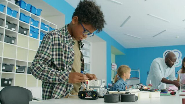 Medium shot of African American boy standing at table in robotics club and constructing toy tractor from plastic blocks, and black male teacher helping two young Caucasian girls in backgroundMedium sh