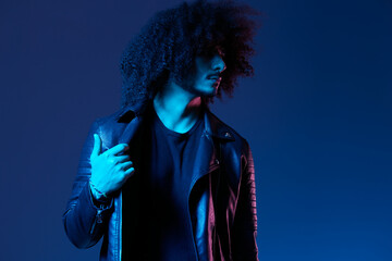 Portrait of fashion man with curly hair on blue background multinational, colored light, black leather jacket trend, modern concept.