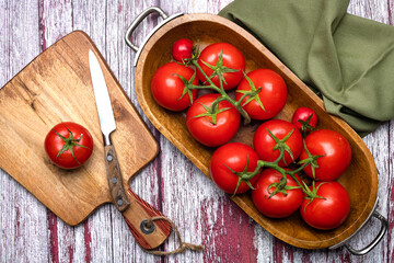 Fresh tomatoes in a wooden bowl and a cutting board with a knife on a wooden table. View from above. Place for text.