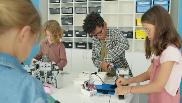 Medium over-shoulder shot of diverse young boys and girls standing around table in classroom and building electric robot models from bright plastic construction kits at after school science club