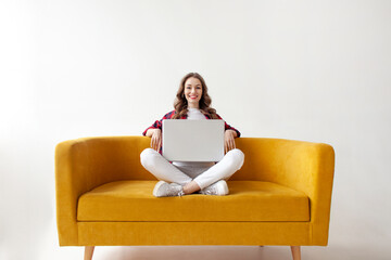 young cute girl uses laptop on comfortable soft sofa, woman types online on computer on yellow couch
