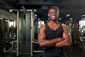 Fototapeta na wymiar young African American man stands with crossed arms in dark gym and smiles, fitness trainer stands in fitness club