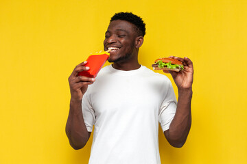 happy african american man in white t-shirt holding big burger and french fries and smiling, guy...