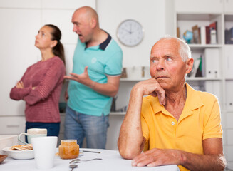 Elderly man sitting at table, unhappy family couple quarrelling on backgroud