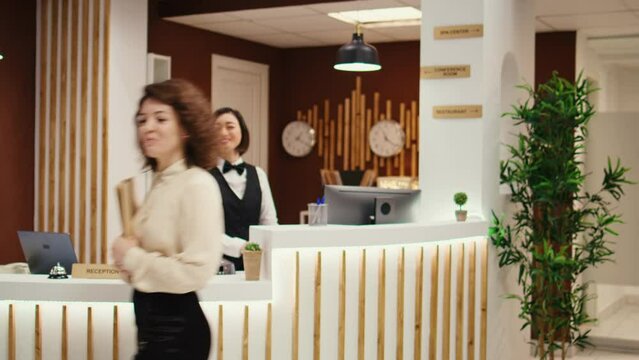 Tracking shot of hotel manager walking towards waiting guests, suggesting hotel extras and room upgrades. Guests discussing booking options and extra requests while being checked in