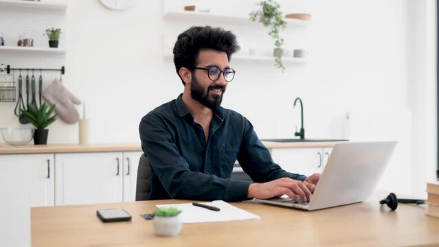 Joyful bearded man in glasses operating modern laptop while sitting at writing desk in spacious dining room. Smiling indian freelancer using flexible schedule while spending working hours at home.