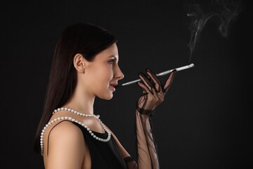 Obraz premium Woman using long cigarette holder for smoking on black background, space for text
