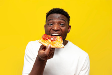 joyful african american man in white t-shirt bites piece of pizza on yellow isolated background,...