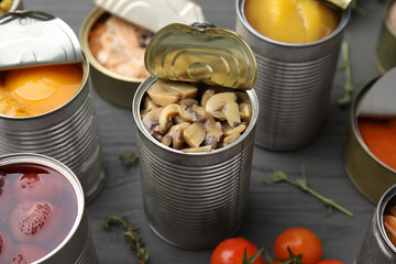 Open tin cans with mushrooms and different products on gray wooden table, closeup