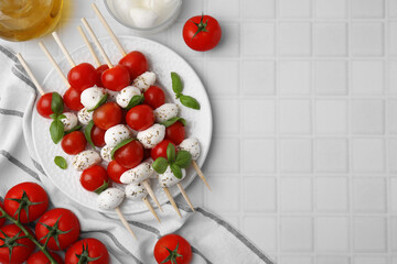 Caprese skewers with tomatoes, mozzarella balls, basil and spices on white tiled table, flat lay. Space for text