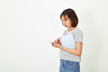 Charming young Asian female employee with short brown hair wearing t-shirt and jeans smiling and holding a digital tablet with smartphone standing on white background copy space