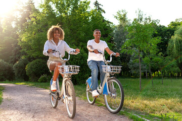 young african american couple riding bike in park and chatting, curly woman and young man are...