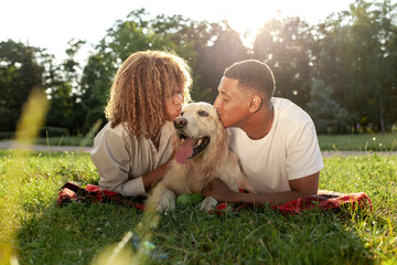 young african american couple with dog lie in park on green grass, curly woman and young man kiss retriever
