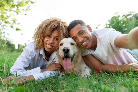 young african american couple with dog lie in park on green grass and take selfie - Powered by Adobe
