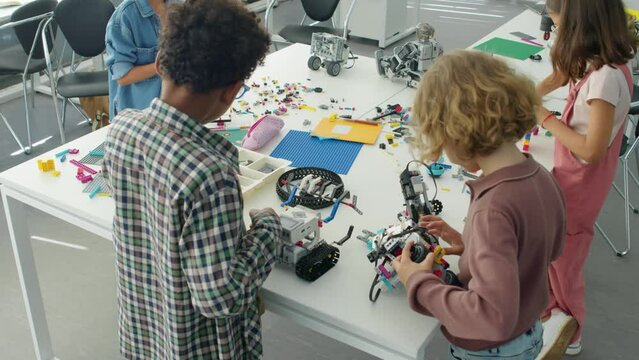 Medium high angle shot of young diverse boys constructing electric robot models on table at after school club, and girls building structures from bright plastic blocks