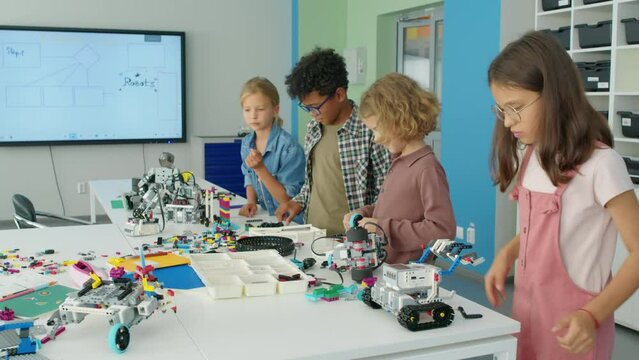 Medium shot of four happy enthusiastic multiracial children standing together at table in hobby club, building toy robot models from plastic construction kits, and chattingMedium shot of four happy en