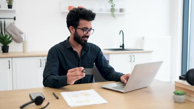 Attractive bearded man in spectacles holding blank credit card while typing on portable computer in kitchen interior. Happy arabian gentleman doing online banking transaction while working remotely.