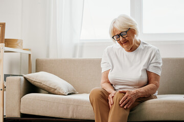 Elderly woman severe pain in her leg sitting on the couch, health problems in old age, poor quality of life. Grandmother with gray hair holds on to her sore knee, problems with joints and ligaments.