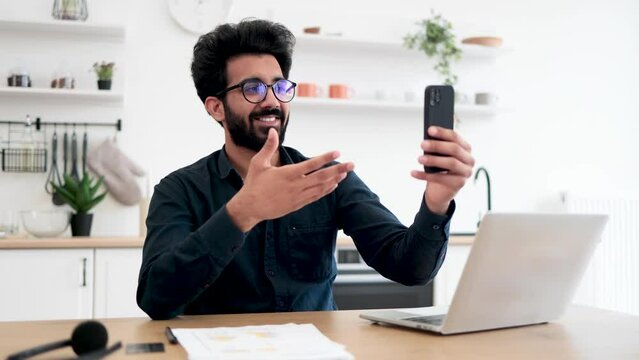 Attractive young man in spectacles holding phone while talking via webcam on kitchen background. Happy indian person in collared shirt conducting online conference via modern device in home interior.