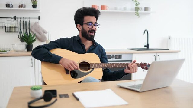 Cheerful arabian person holding string instrument while smiling at portable computer webcam in spacious apartment. Happy bespectacled male teaching online guitar lesson using modern technologies.