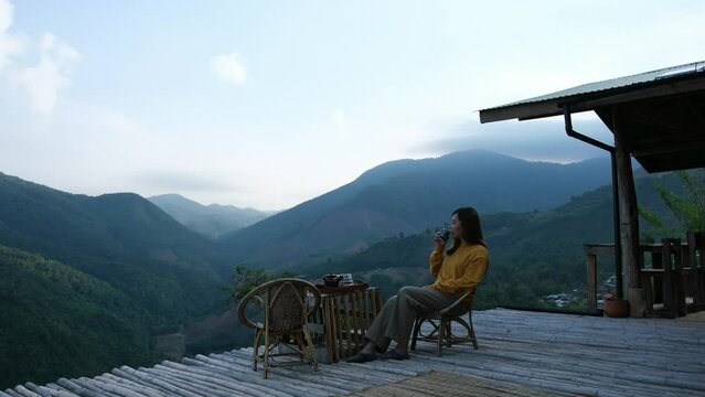 A female traveler drinking hot coffee while sitting on wooden terrace with a beautiful mountains view