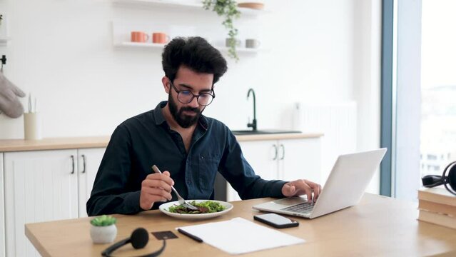 Efficient indian worker in glasses eating vegetables while typing on portable computer at wooden desk in kitchen. Remote freelancer performing business task without wasting time for lunch break.