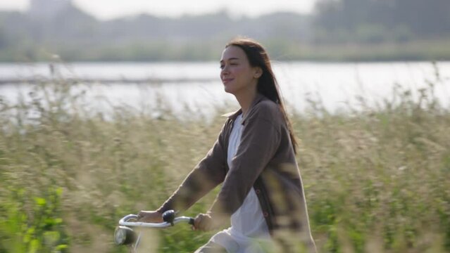 Smiling happy woman cycles along river bank with wind through her hair, tracking