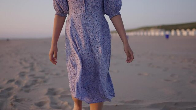 Smiling brunette in flowy summer dress walks barefoot on beach. Tracking back