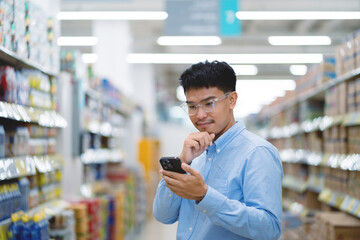 Asian man shopping for groceries, calculating product prices, looking at shelves with food products standing in supermarket.