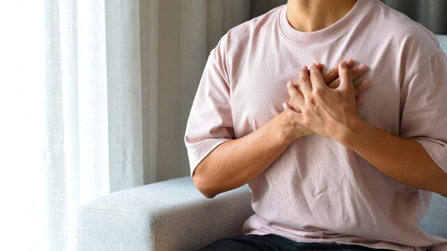 Young man sitting on sofa in the living room using both hands holding chest with symptom heart attack disease.He was worried because of the pain on the left side of his chest.