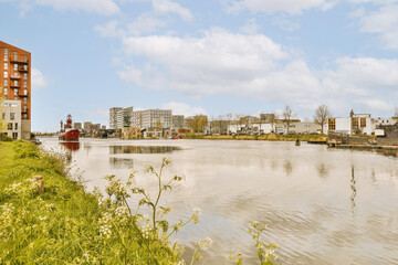 a river with buildings in the background and green grass growing along the water's edge, on a sunny...