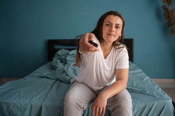 Woman watches TV sitting on the bed, using tv remote control in her hands. Morning or evening in the bedroom