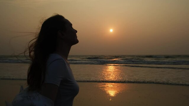 Happy long-haired woman traveller enjoys sea breeze on the seashore at sunset. Female profile silhouette with flowing hair on the setting sun background.