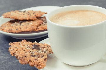 Cup of white coffee and fresh baked oatmeal cookies with honey and healthy seeds on plate. Delicious crunchy dessert