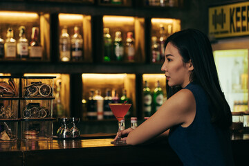 Asian woman drinking a cocktail at a bar at night.