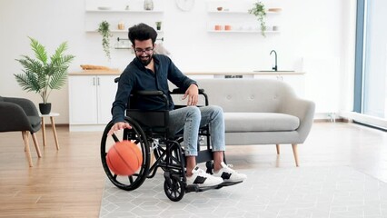 Cheerful bespectacled adult with mobility impairment dribbling ball in studio apartment on weekend. Happy indian in jeans and dark shirt staying active with disability while playing game for pleasure.