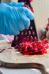 Person, wearing rubber gloves, grating fresh raw beetroot with a metal grater in a kitchen