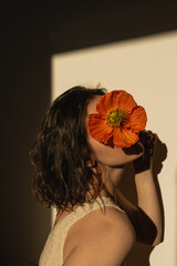 Young pretty woman hide face with red poppy flower over tan beige wall with aesthetic sunlight...