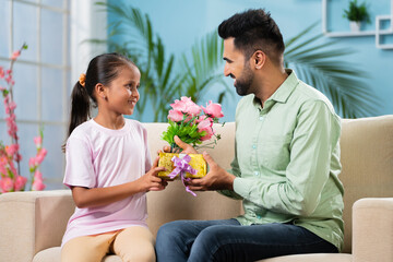 Happy young Indian girl kid giving flowers with gift to father while sitting on sofa at home - concept of father's day, relationship and family affection.