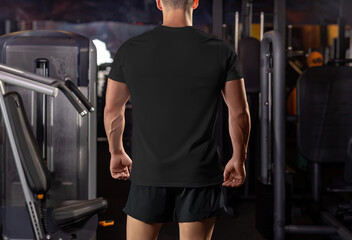Mockup of a black t-shirt on a muscular guy against the background of equipment in the gym, rear...