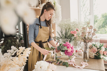 woman florist in beige apron is working in  flower shop. Floral design studio, making decorations and arrangements. Flower delivery, order creation. Small business. - obrazy, fototapety, plakaty