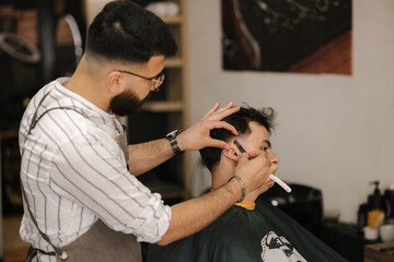 Side view of barber using sharp blade for shaving. Handsome customer sitting in barbershop