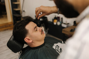 Process of threading procedure in barber shop. Professional barber correcting shape of brows with threads male client sitting in chair.